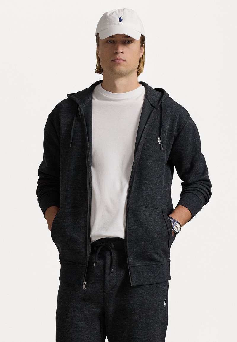 Sweatshirt preta com fecho, com um bolso frontal, feita de um material suave. Combinada com uma t-shirt branca e calças de fato escuras. Logótipo icónico no canto superior esquerdo.