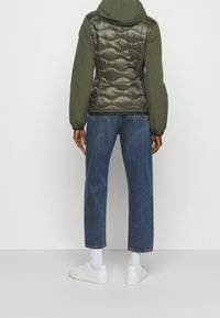 Olijfgroene gewatteerde jas met capuchon, met een getextureerde voorkant en gladde mouwen. Gecombineerd met losse blauwe denim jeans en witte sneakers.