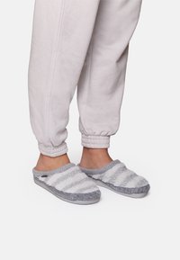 Grå och vita randiga tofflor med en mjuk och fluffig textur. Öppen bak och rundad tån design. Bärs med grå uppvikta sweatpants.