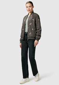 Kvinde iført en grå bomberjakke med blomsterprint, lysegrå top, sorte jeans og hvide sneakers, stående foran en ensfarvet lys baggrund.