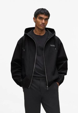 Joven con una chaqueta con capucha negra con cremallera sobre una camiseta negra y pantalones negros, de pie con las manos en los bolsillos frente a un fondo blanco.