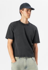 Tricou scurt cu mânecă scurtă, de culoare gri închis, cu guler rotund și croială lejeră; material moale cu o textură ușor purtată. Purtați cu blugi de denim albastru deschis.