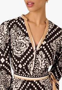 Schwarzes Kleid mit weißen Blumen- und Polka-Dot-Mustern, verziert mit einem beigen Saum, V-Ausschnitt und einem gebundenen Gürtel, der die Taille betont.