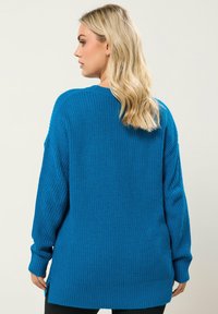 Pull en tricot bleu avec une coupe ample, manches longues, texture côtelée et fentes sur les côtés pour faciliter le mouvement. Ourlet évasé et col rond.