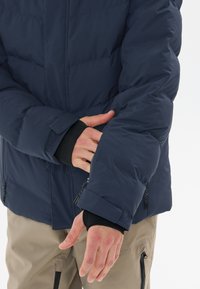 Marineblaue Steppjacke mit einem Reißverschluss an den Bündchen und einem Klettverschluss, gestepptem Design und glatter Textur, getragen über einer schwarzen Schicht.