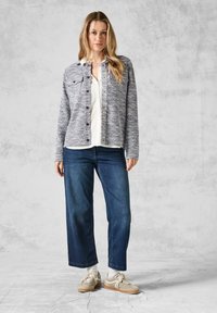 Texturerad marinblå jacka med ljus rutigt mönster, knäppning framtill, parat med blå vida jeans och beige sneakers. Vit skjorta under.