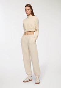 Giacca beige cropped con zip e colletto alto, abbinata a pantaloni della tuta larghi e coordinati. Sneakers bianche con dettagli neri completano il look.