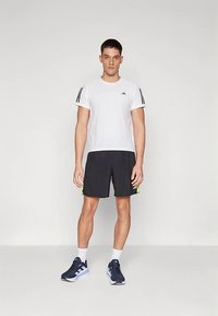 Camiseta deportiva blanca de manga corta con rayas negras, combinada con pantalones cortos negros con un detalle verde y zapatos deportivos oscuros.