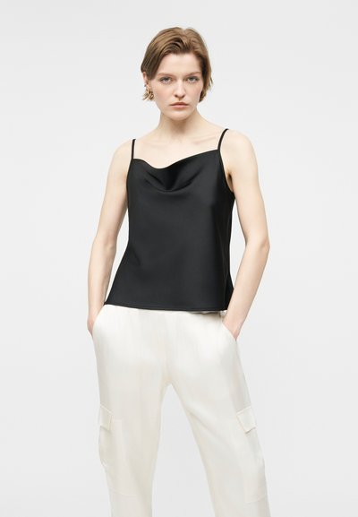 Γυναίκα με κοντά μαλλιά, φορώντας μαύρο σατέν μπλουζάκι χωρίς μανίκια και άνετο λευκό παντελόνι με τα χέρια στις τσέπες, μπροστά σε απλό φόντο.