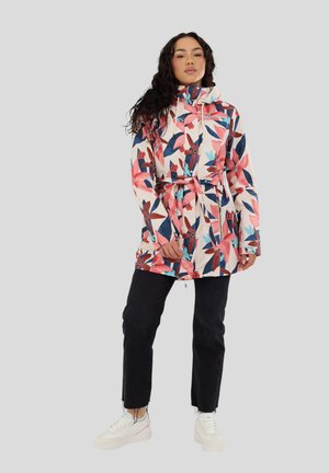 Frau trägt eine floral gemusterte, taillierte Jacke mit Kapuze, schwarze Hose und weiße Sneakers und steht vor einem einfarbigen grauen Hintergrund.