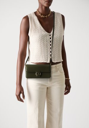 Grønn lær crossbody-veske med klaffelukking, gullfarget maskinvare og avtakbar stropp. Modellen har på seg en kremfarget strikket vest og lyse bukser.