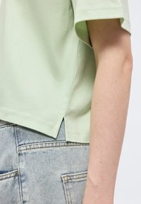 Ανοιχτό πράσινο πουκάμισο με κοντά μανίκια και πλαϊνή σχισμή, φορεμένο με τζιν παντελόνι σε ανοιχτό μπλε, που δείχνει το χέρι και τη μέση ενός ατόμου.