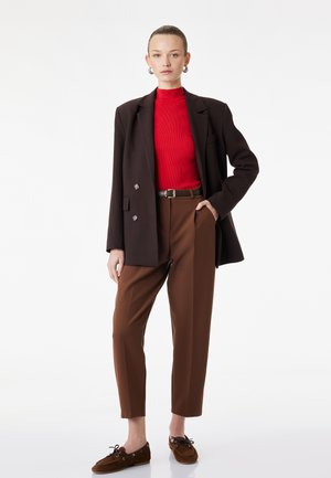 Vrouw staand, dragend een rood topje, donkerbruine blazer, bruine hoge taille broek, bruine riem, bruine loafers en zilveren oorringen.