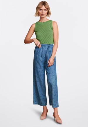 Groen mouwloos topje met een gestructureerd patroon, gecombineerd met brede blauwe broeken met een subtiele golvende print. Het model draagt lichtroze hakken.