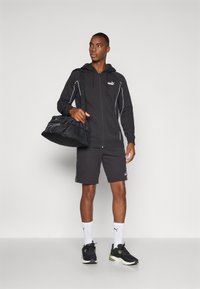 Svart zip-up hettegenser med hvite detaljer, svarte shorts, svarte joggesko og hvite sokker med logo, holder en svart treningsbag.