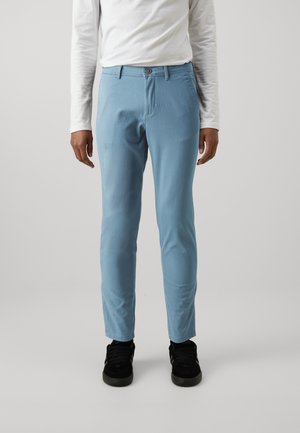 Pantalones azul claro de corte recto, con bolsillos frontales y cierre de botón, combinados con zapatos negros y una camisa blanca de manga larga.