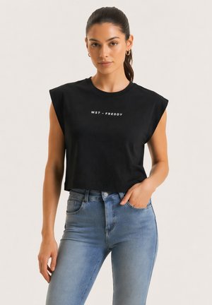 Nainen, jolla on päällään musta hihaton lyhyt toppi, jossa teksti "MST - FREDDY" ja siniset korkeavyötäröiset farkut, seisoo toinen käsi taskussa.