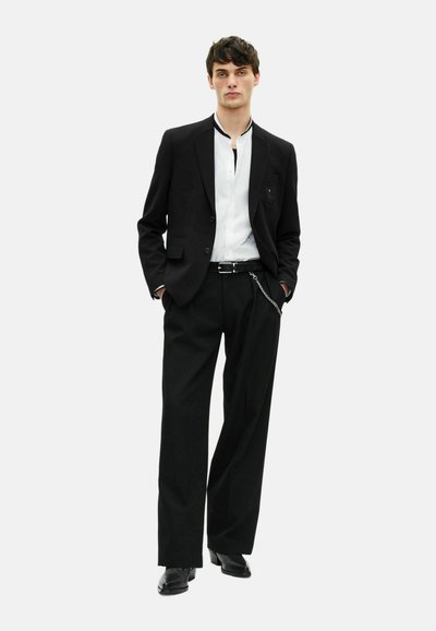Schwarzer Anzug mit einem taillierten Blazer über einem weißen Hemd. Die Hose ist locker geschnitten und hat einen Reißverschluss mit Kettendetails. Schwarze Schuhe vervollständigen den Look.