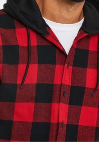 Man in een rood en zwart geruit overhemd met knopen, met een zwarte capuchon en een witte onderhemd, afgekapt onder de hals.