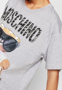 Grå bomullströja med en grafik av en björn som bär solglasögon, med fet svart "Moschino"-text längst upp. Korta ärmar.