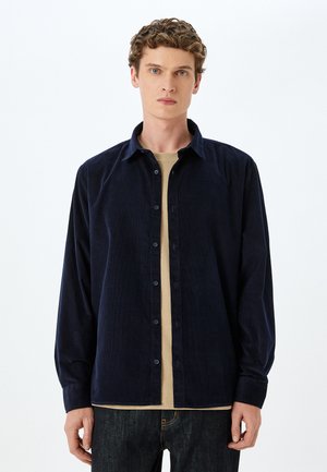 Marineblå kordfløjls skjorte med lange ærmer, der har en klassisk krave og knaplukning foran, lagdelt over en lys beige top.