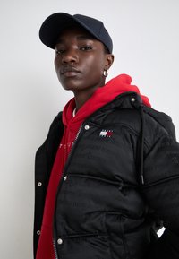 Black puffer jacket with a glossy finish and tonal logo print, paired with a red hoodie, navy cap, and silver hoop earring.