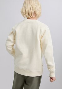 Hanorac crem cu textură moale, mâneci raglan, guler rotund și manșete ribbed. Purtat cu pantaloni verde măsliniu.