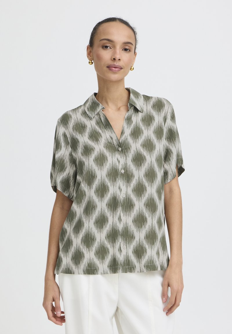 Nő zöld-fehér mintás, rövid ujjú, gombos blúzban és fehér nadrágban, egyszínű háttér előtt áll.