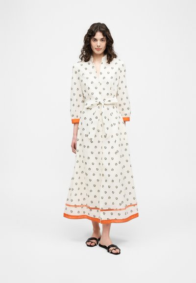 Femme aux cheveux bouclés portant une robe longue blanche à petites imprimés floraux, avec des poignets et un ourlet orange, une taille nouée, et des sandales noires, debout sur un fond blanc.