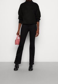 Pull noir en maille avec col montant, associé à un jean noir ajusté avec des boutons décoratifs à l'ourlet, et des bottes noires à talons. Tenant un sac rose.