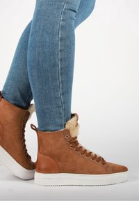 Ruskea mokkanahkainen korkea sneakeri, jossa on keinoturkisvuori, metalliset aukot, pyöreät nauhat ja valkoinen kumipohja.