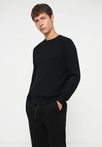 Schwarzer, gerippter Strickpullover mit Rundhalsausschnitt und langen Ärmeln, kombiniert mit schwarzen Jogginghosen mit Zugband. Das Material wirkt weich und strukturiert.