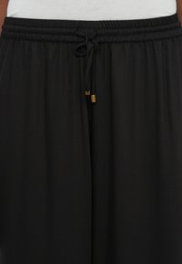 Detailný záber na čierne nohavice s elastickým pásom a viazacou šnúrkou, ktorá má zlaté kovové konce, s bielym vrchom čiastočne viditeľným nad pásom.