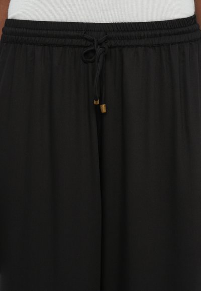 Close-up of black elastic-waist pants with a tied drawstring featuring gold metal tips and white top partially visible above waistband.