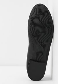 Semelle de chaussure habillée noire avec motif de bande de roulement ondulé texturé, présentée sur fond blanc, mettant en avant le design antidérapant.