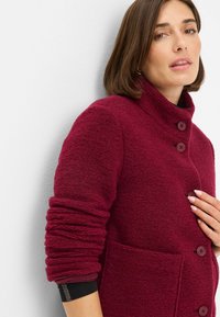 Cappotto in misto lana borgogna con colletto alto, chiusura a quattro bottoni e due tasche frontali. Tessuto strutturato con maniche arrotolate.