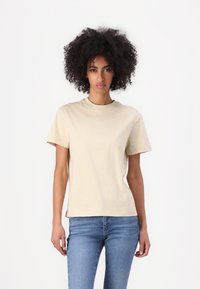 Beige cotton t-shirt with a round neckline and short sleeves, featured on a person wearing blue jeans, set against a white background.