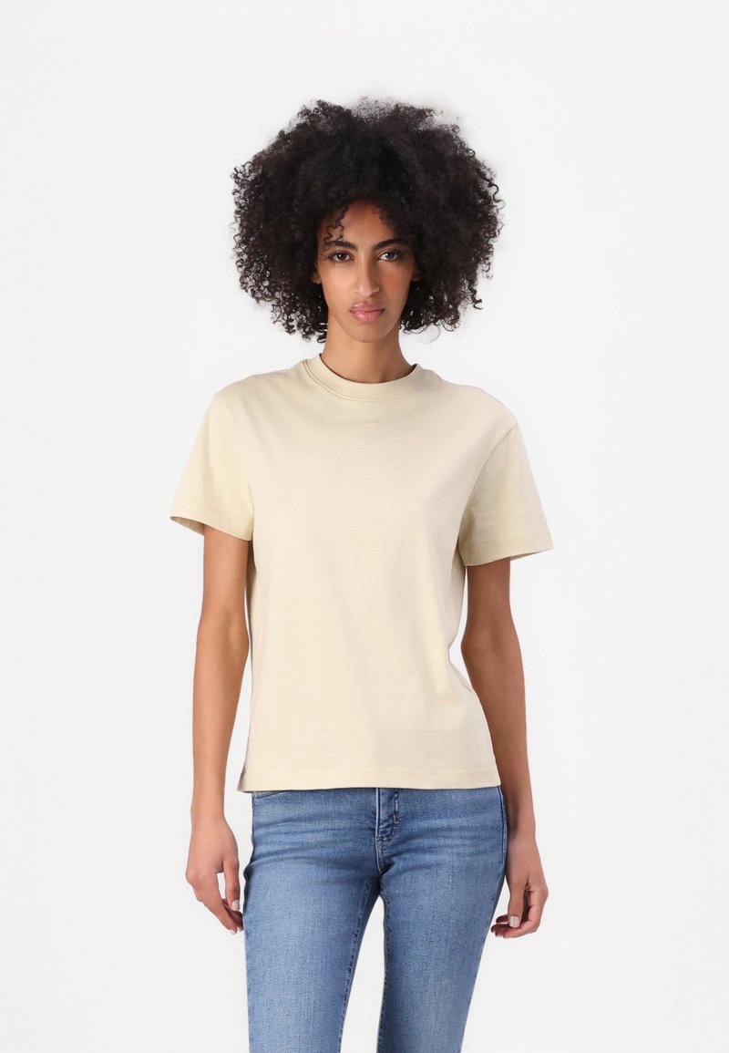 Beige cotton t-shirt with a round neckline and short sleeves, featured on a person wearing blue jeans, set against a white background.