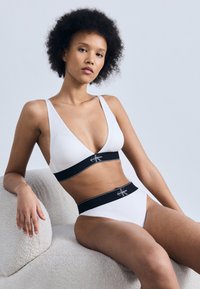 Γυναίκα με σγουρά μαλλιά, φορώντας λευκά εσώρουχα Calvin Klein με μαύρες ελαστικές ταινίες, καθισμένη σε μια υφασμάτινη ανοιχτόχρωμη καρέκλα μπροστά σε μονόχρωμο φόντο.