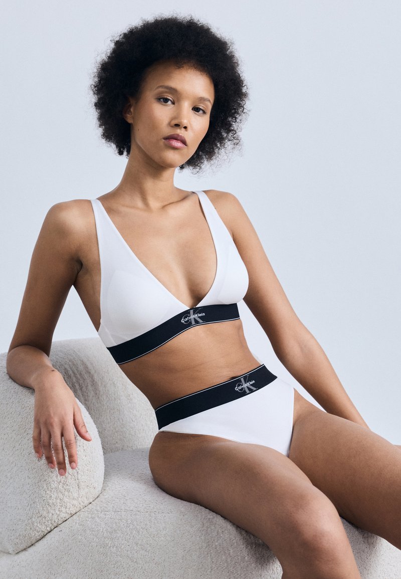 Γυναίκα με σγουρά μαλλιά, φορώντας λευκά εσώρουχα Calvin Klein με μαύρες ελαστικές ταινίες, καθισμένη σε μια υφασμάτινη ανοιχτόχρωμη καρέκλα μπροστά σε μονόχρωμο φόντο.