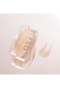Klar glasburk med skruvlock, innehållande en ljusbeige kräm. Etiketten lyder "OSKIA LONDON." En liten mängd är utstruken på en yta.