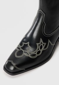 Bota de vaquero de cuero negro con punta afilada, decorada con costuras blancas y bordado floral a lo largo del costado, acabado de textura suave.