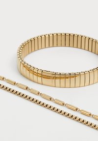 Smykke sett i gulltone som inkluderer et fleksibelt armbånd med rektangulære lenker og to kjeder: en med vekslende lenker og en glatt overflate.