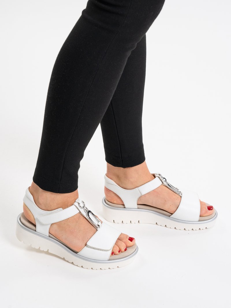 Pies con sandalias blancas de plataforma con detalles de hebilla plateada y leggings negros sobre un fondo blanco.