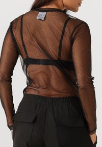 Top lungo a maniche nere in mesh trasparente con motivo a pois. Presenta un cinturino nero visibile e un'etichetta sul retro, abbinato a pantaloni con cintura elastica nera.
