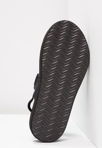 Semelle de chaussure noire avec motif ondulé reposant contre un coin de mur et de sol blanc, montrant la texture de l'adhérence.