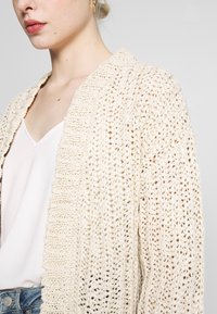 Gebreide cardigan in beige, met een losse weefstructuur, dikke textuur en brede revers. Gecombineerd met een lichte cami, met zichtbare spijkerbroek.