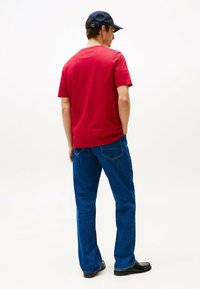 Rød bomulds t-shirt med korte ærmer, kombineret med blå denim jeans. Modellen bærer en marineblå hat og sorte sko.