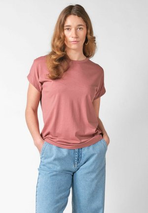 Žena s dugom valovitom kosom, odjevena u ružičastu majicu kratkih rukava i svijetloplave traperice, s rukama u džepovima, stoji nasuprot jednostavnoj pozadini.