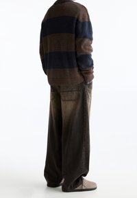 Personne debout, les mains dans les poches, portant un pull rayé marron et bleu marine, un jean large foncé et des chaussures en daim beige.